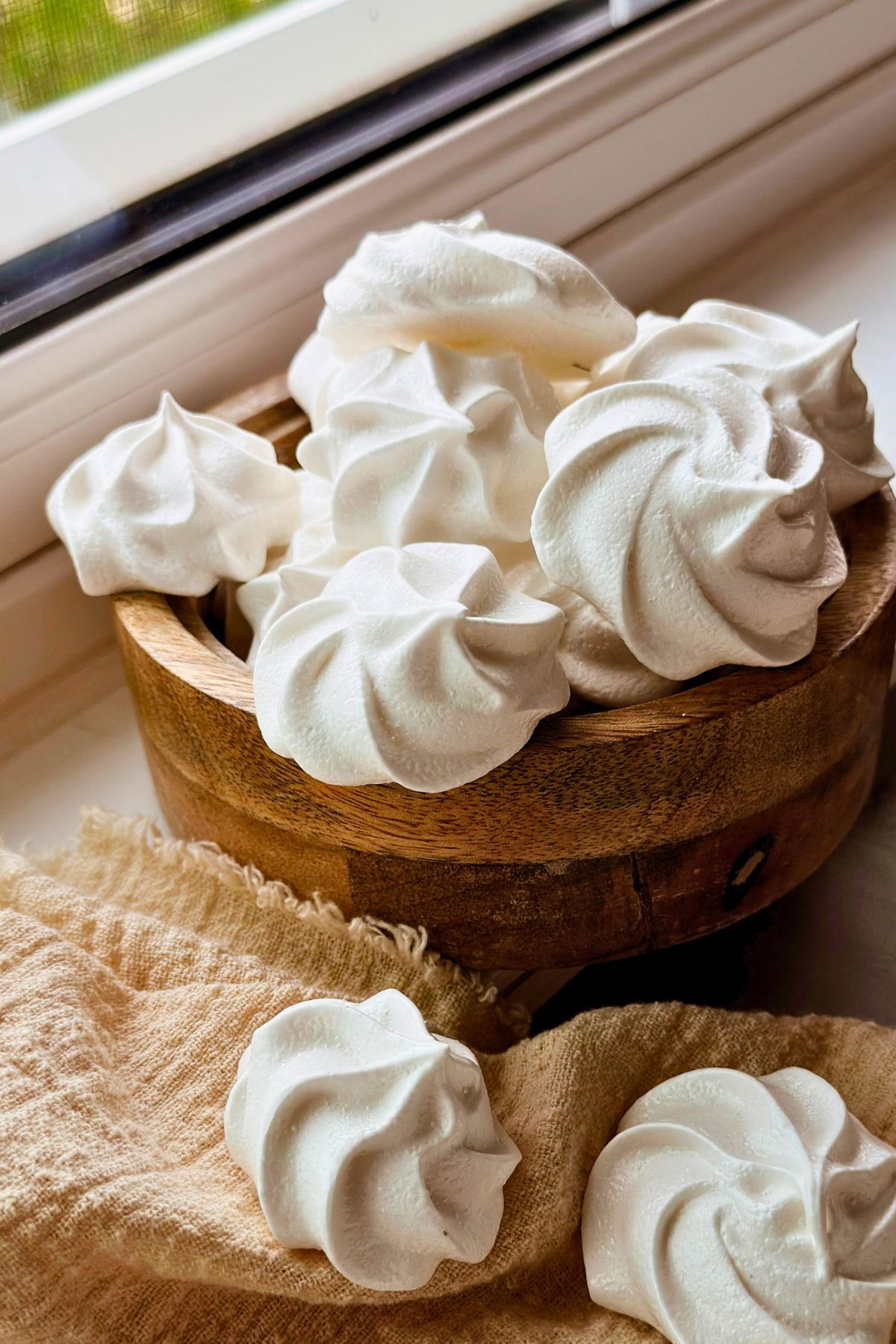 A rustic wooden bowl overflowing with white Cuban suspiros meringue cookies, styled on a warm beige linen cloth near a sunny window. Two suspiros rest on the cloth in the foreground, showing their swirled rosette shape and crisp, airy texture.