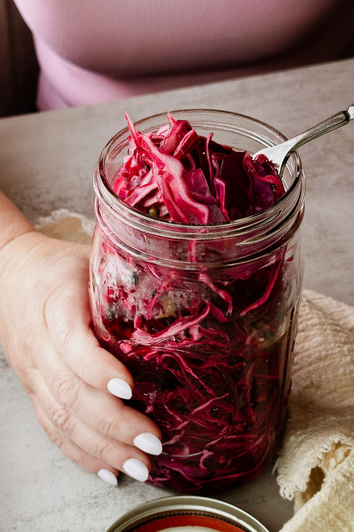 A hand with white nails using a fork to lift a garlic clove from a large mason jar packed with deep red pickled cabbage, photographed near a window with soft natural light and a yellow linen napkin beside it.