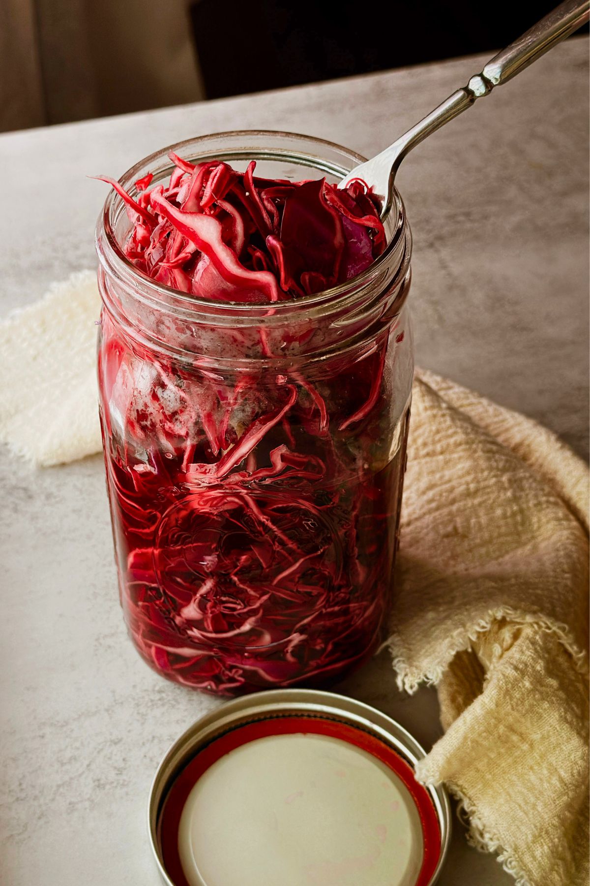 Image 1:
A quart mason jar filled with bright red pickled cabbage and a fork resting inside, with the lid off and a yellow linen napkin alongside it on a light stone surface.