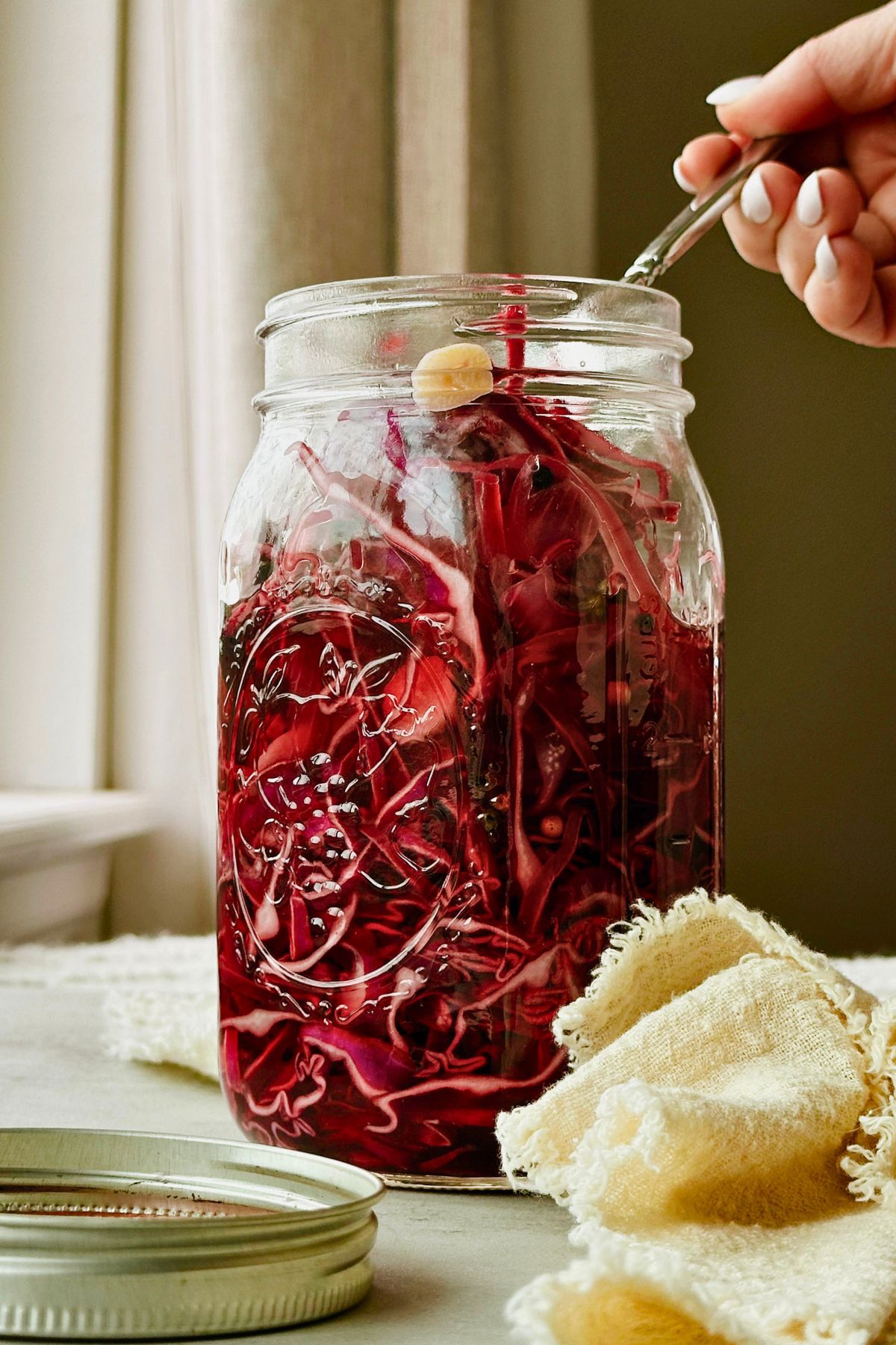 A hand with white nails holding a quart mason jar of pickled red cabbage with a fork inside, against a light gray surface with a yellow linen napkin nearby.
