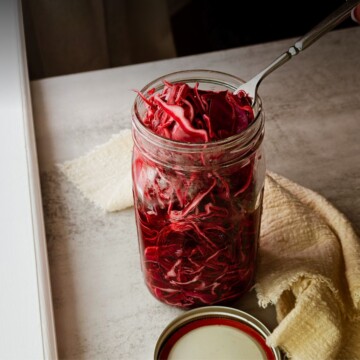 A quart mason jar packed with bright red pickled cabbage and a fork resting inside, with the lid set aside and white and yellow linen napkins on a light stone surface.