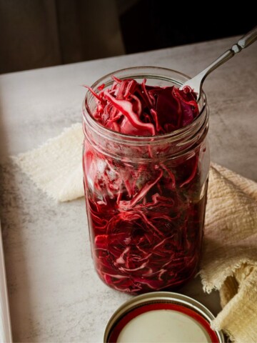 A quart mason jar packed with bright red pickled cabbage and a fork resting inside, with the lid set aside and white and yellow linen napkins on a light stone surface.