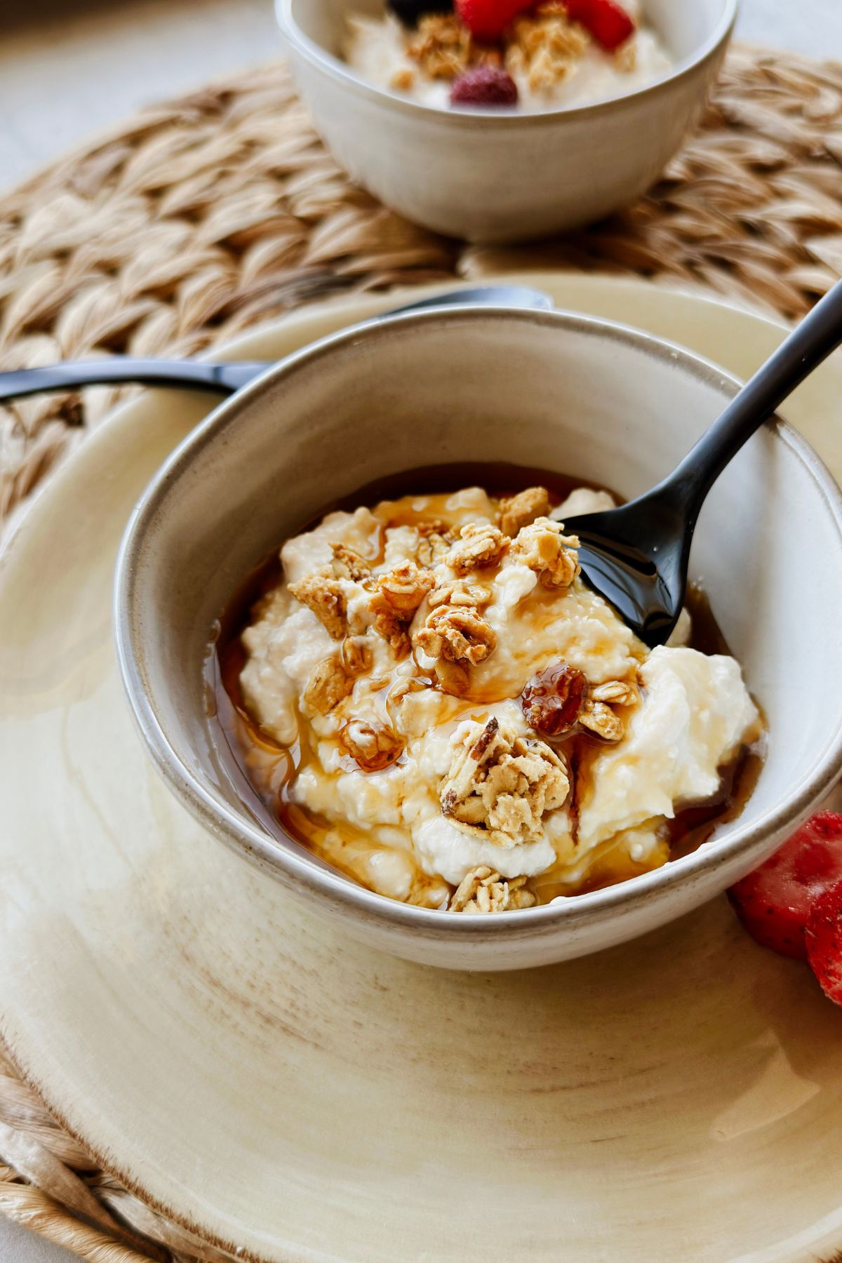 A bowl of thick, creamy vegan Greek yogurt drizzled with golden maple syrup or agave and topped with crunchy granola, served in a rustic ceramic bowl on a yellow plate with a black spoon, fresh strawberries and blueberries on the side.