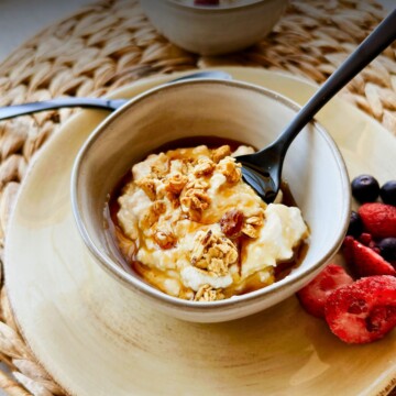 A bowl of thick, creamy vegan Greek yogurt drizzled with golden maple syrup or agave and topped with crunchy granola, served in a rustic ceramic bowl on a yellow plate with a black spoon, fresh strawberries and blueberries on the side.