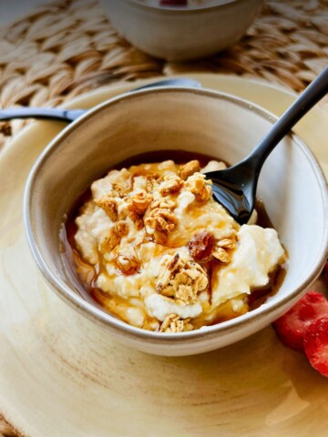 A bowl of thick, creamy vegan Greek yogurt drizzled with golden maple syrup or agave and topped with crunchy granola, served in a rustic ceramic bowl on a yellow plate with a black spoon, fresh strawberries and blueberries on the side.
