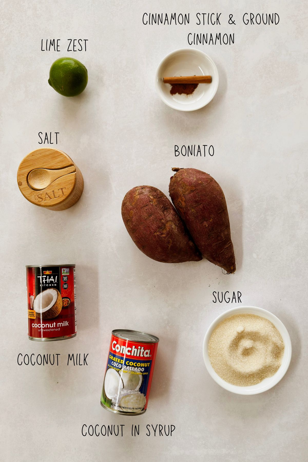 Ingredients for Boniatillo Con Coco laid out on a table: boniato, canned coconut milk, grated coconut in syrup, sugar, a cinnamon stick with ground cinnamon, a lime, and salt.