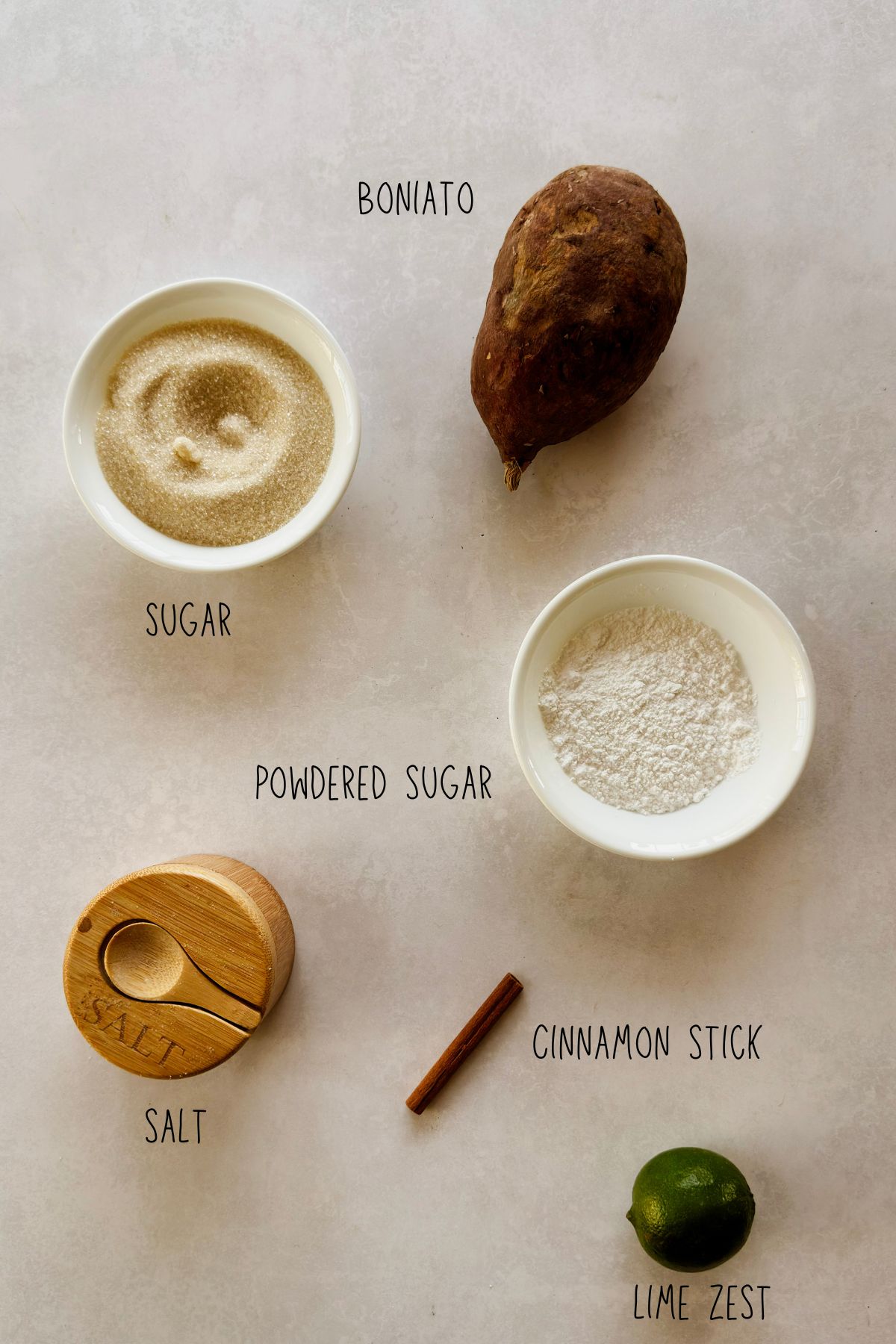 Ingredients for boniatillo seco on a light stone surface: whole boniato, granulated sugar, powdered sugar, salt, a cinnamon stick, and a lime.