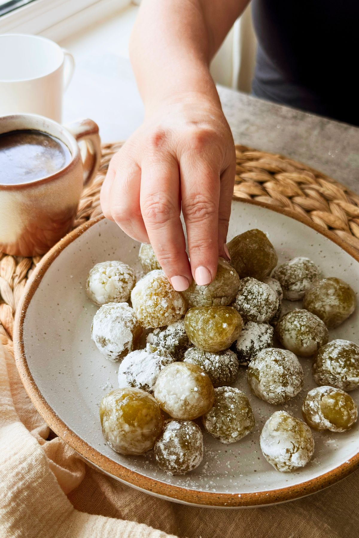 Hand reaching for a boniatillo seco candy from a plate of powdered sugar-coated Cuban sweet potato balls beside a mug of coffee.