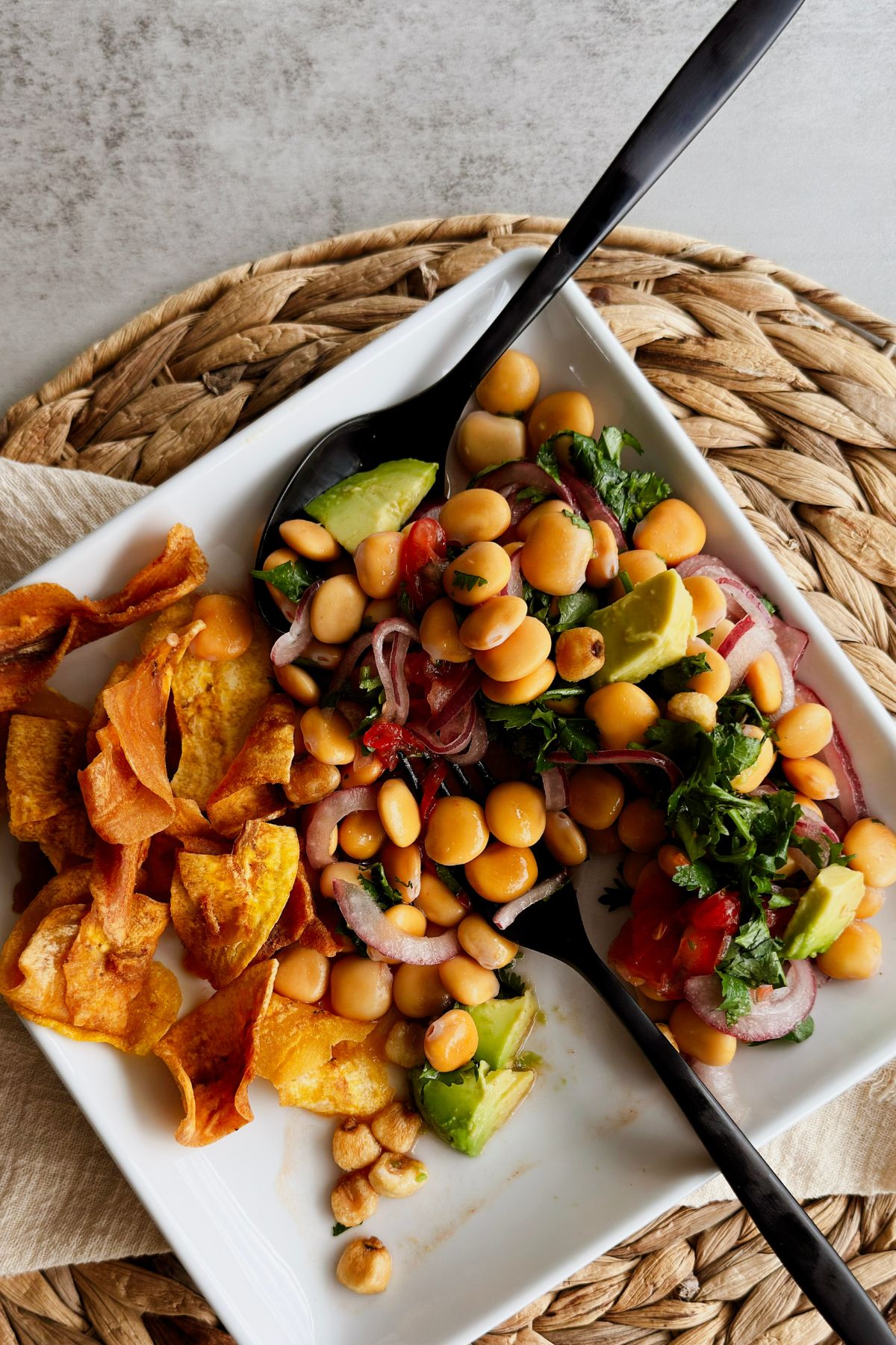 Ceviche de chochos served on a square white platter with a black serving spoon, lupini beans with red onion, cilantro, tomato, avocado, and corn nuts beside a pile of plantain chips.