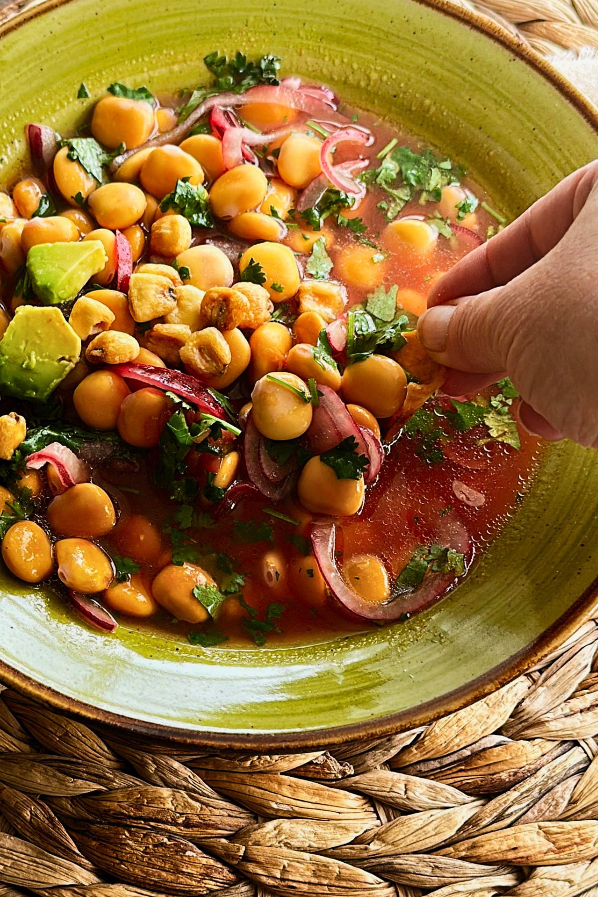 A hand scooping ceviche de chochos with a plantain chip from a green ceramic bowl, lupini beans and red onion in a coral citrus marinade with corn nuts, cilantro, and avocado.