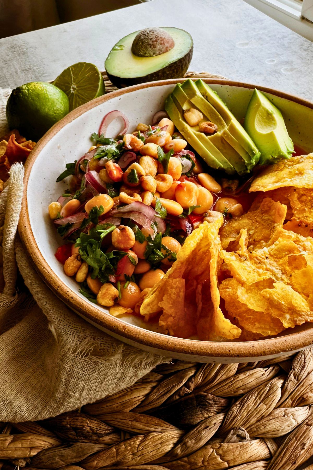 Ceviche de chochos in a speckled ceramic bowl with lupini beans, red onion, cilantro, corn nuts, sliced avocado, and crispy plantain chips, with a lime and half an avocado behind it.