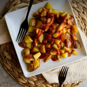Patatas bravas on a square white plate, crispy potato chunks drizzled with smoky red bravas sauce, two black forks alongside.