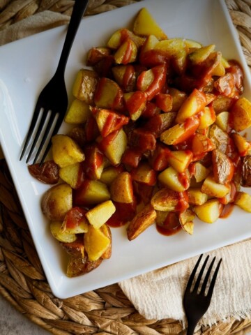 Patatas bravas on a square white plate, crispy potato chunks drizzled with smoky red bravas sauce, two black forks alongside.