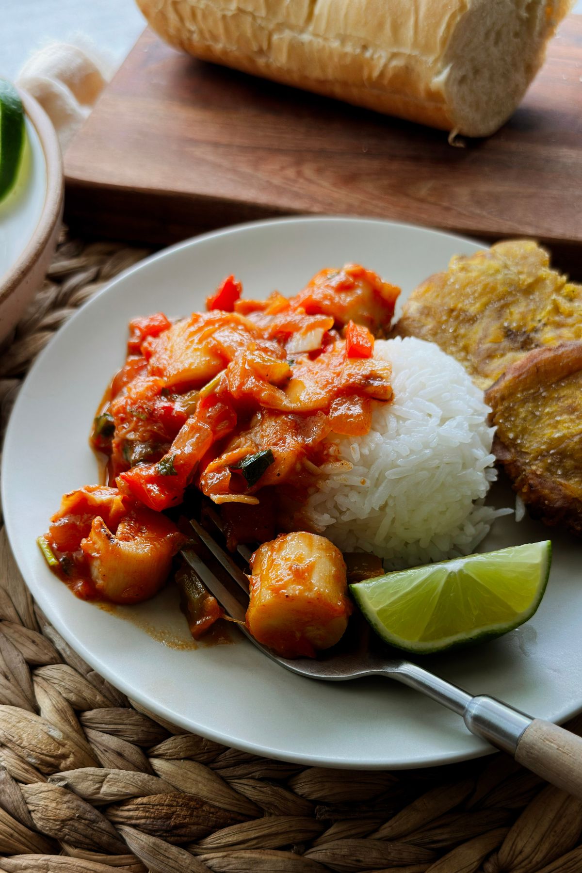 Close-up of vegan camarones enchilados on a plate with white rice, tostones, and a lime wedge, a forkful of hearts of palm in front and Cuban bread behind.