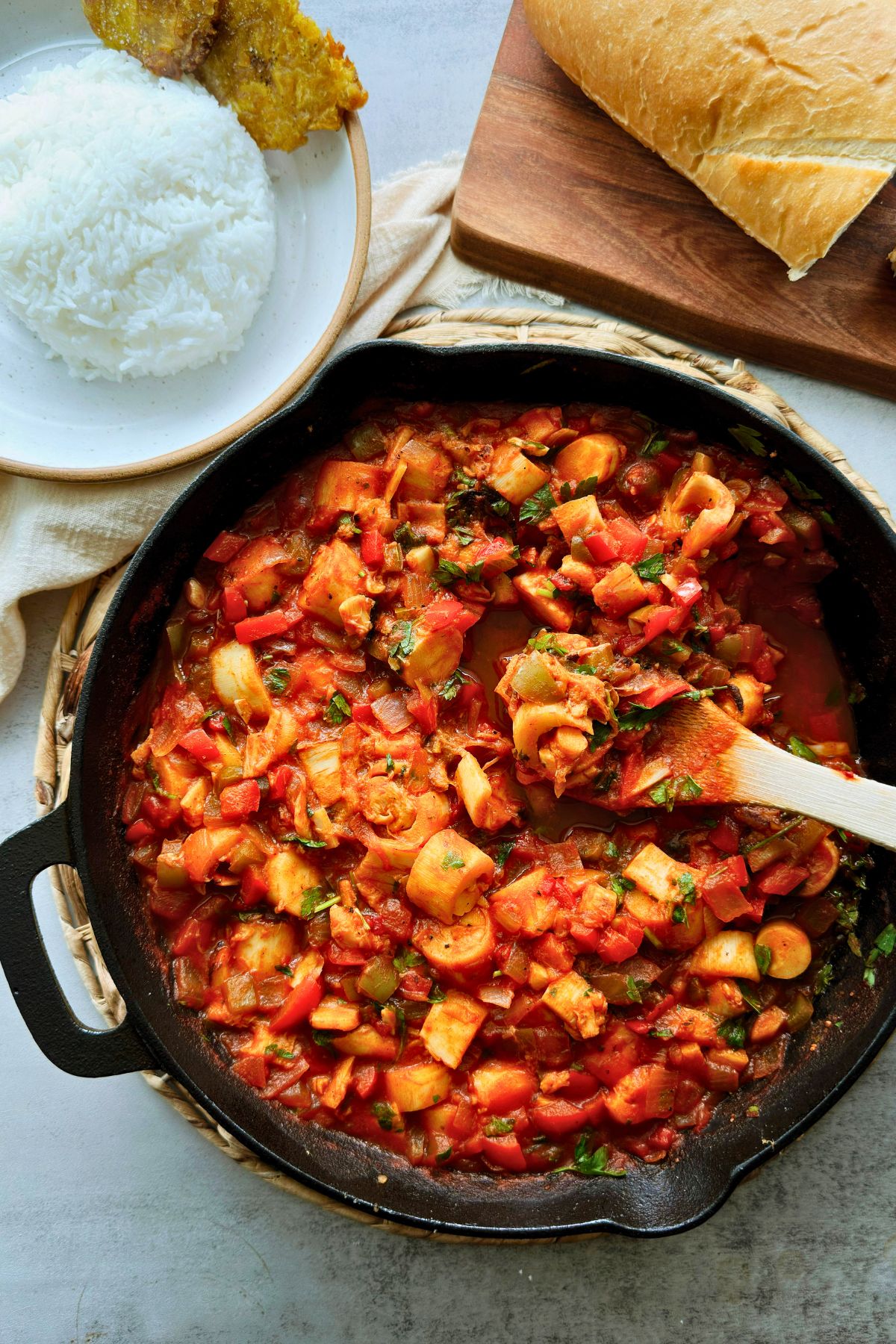 Overhead view of a cast iron skillet of vegan camarones enchilados with a wooden spoon, plates of white rice and tostones and a loaf of bread alongside.