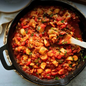 Close overhead shot of vegan camarones enchilados in a cast iron skillet, hearts of palm and diced peppers in red sauce with a wooden spoon resting in the pan.