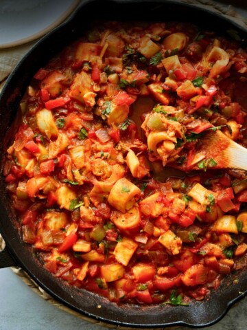 Close overhead shot of vegan camarones enchilados in a cast iron skillet, hearts of palm and diced peppers in red sauce with a wooden spoon resting in the pan.