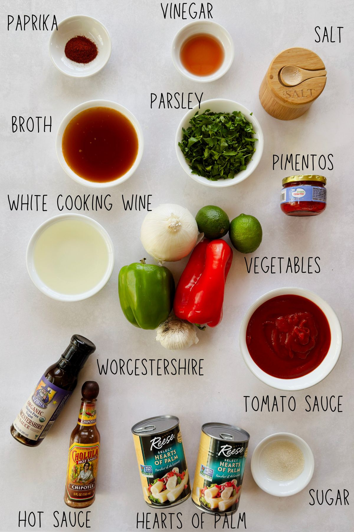 Ingredients for vegan camarones enchilados labeled on a white surface, including hearts of palm, tomato sauce, bell peppers, onion, garlic, limes, pimentos, and vegan Worcestershire sauce.