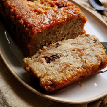 Thick slice of vegan Cuban pudín de pan with visible raisins and custardy bread layers, resting against the caramel-topped loaf on an oval platter.