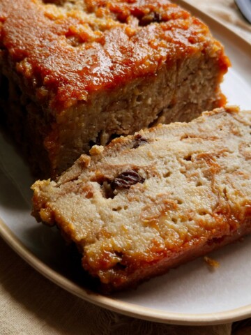 Thick slice of vegan Cuban pudín de pan with visible raisins and custardy bread layers, resting against the caramel-topped loaf on an oval platter.