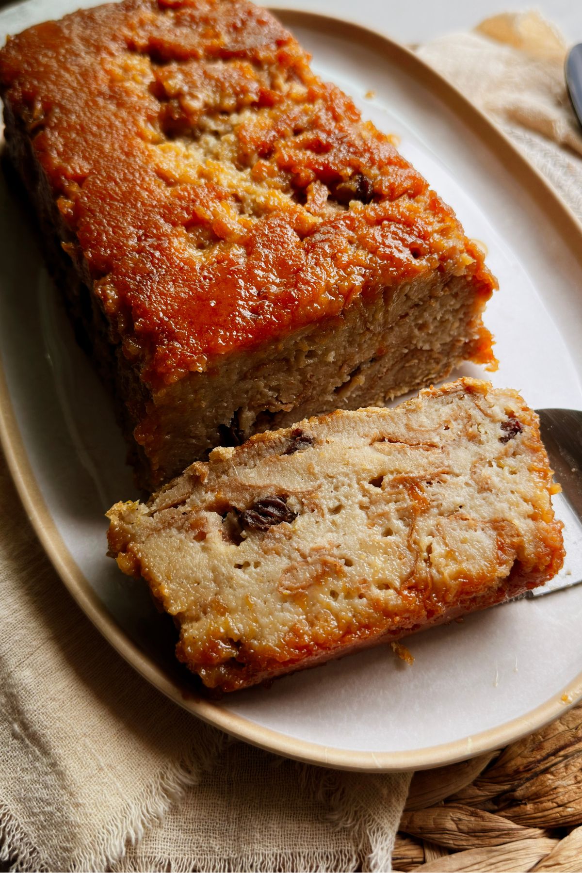 Sliced vegan pudín de pan on an oval platter, one raisin-studded slice lifted on a knife from the caramel-topped loaf.