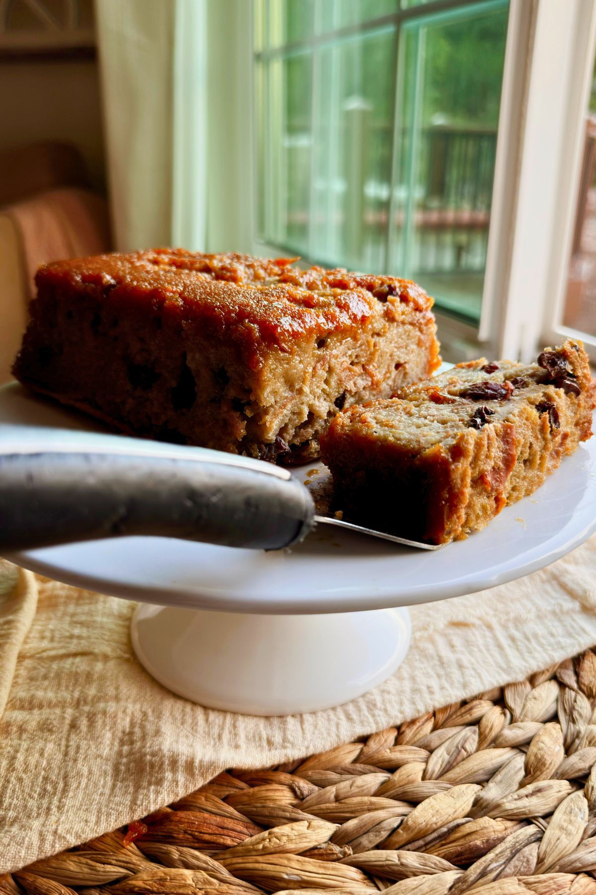 Close-up of a slice of vegan pudín de pan showing raisins and soft bread layers, cut from the caramel-topped loaf on a plate by a sunny window.