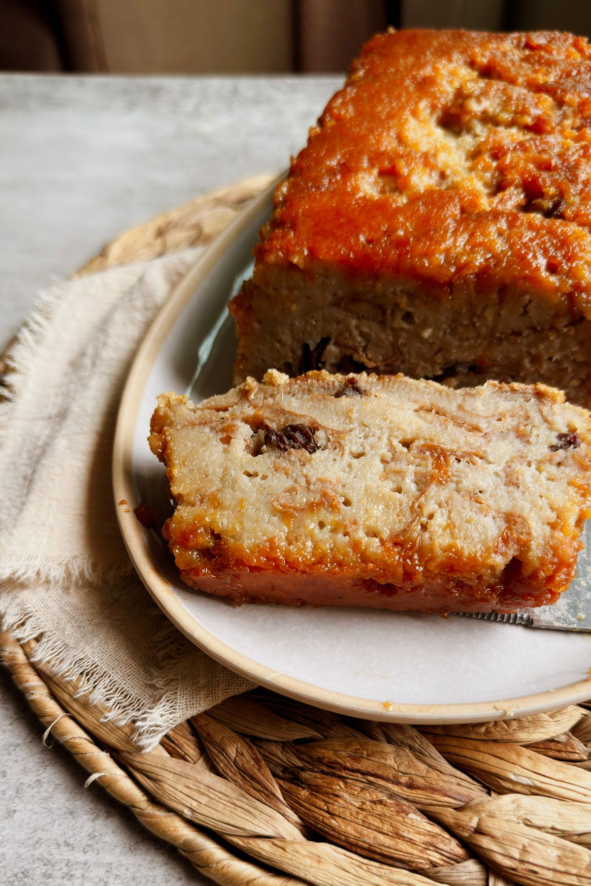 A slice of vegan pudín de pan with a caramel-glazed top and raisins, resting on a ceramic plate in front of the rest of the loaf.
