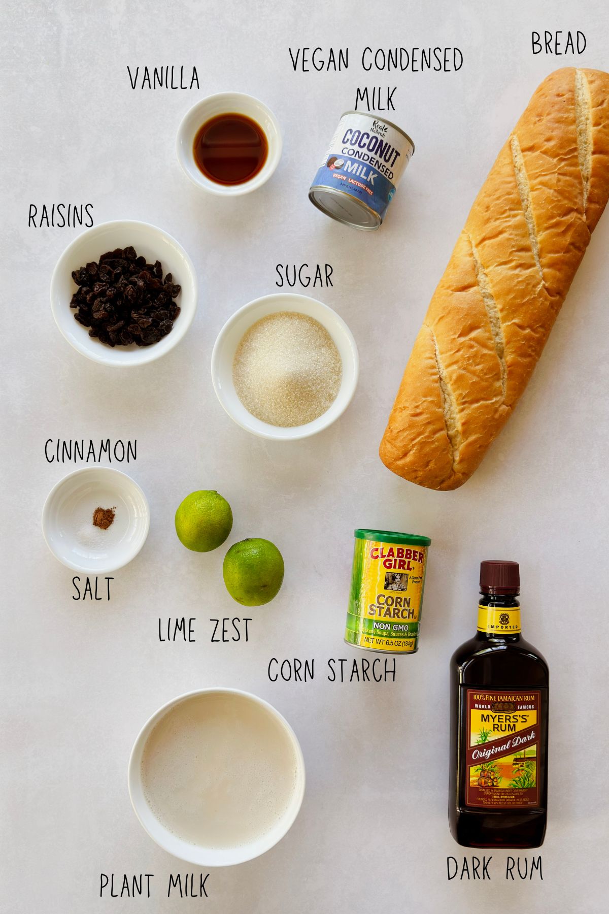 Labeled ingredients for vegan Cuban pudín de pan: bread, coconut condensed milk, plant milk, dark rum, raisins, limes, cornstarch, sugar, vanilla, and cinnamon.