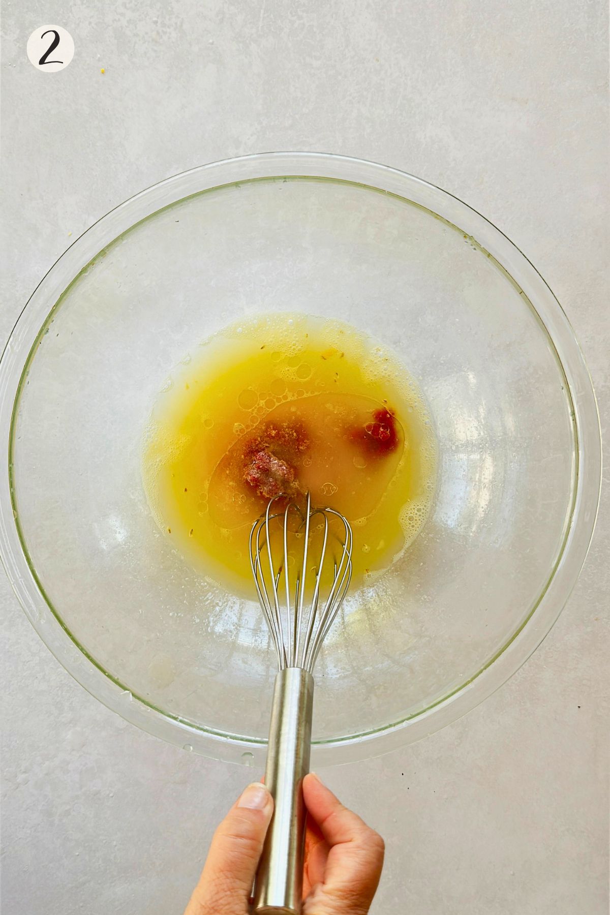 Step 2 of making ceviche de chochos: a hand whisking orange juice, lime juice, salt, oil, and tomato paste together in a clear glass bowl, the paste not yet dissolved.
