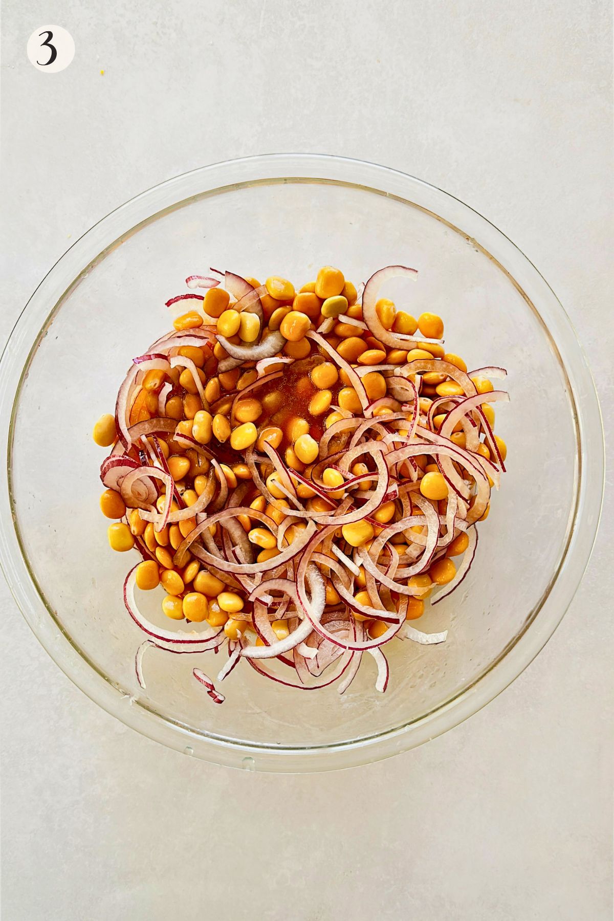 Step 3 of making ceviche de chochos: drained lupini beans and sliced red onion added to the citrus and tomato paste marinade in a glass bowl.