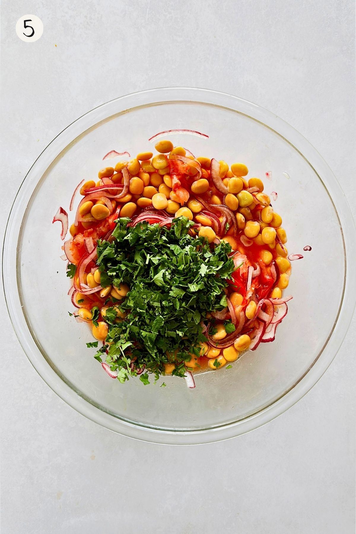 Step 5 of making ceviche de chochos: a mound of freshly chopped cilantro added to the marinated lupini beans, red onion, and tomato in a glass bowl.