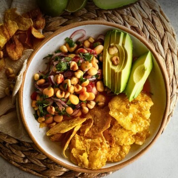Overhead view of ceviche de chochos in a speckled bowl with lupini beans, red onion, cilantro, and corn nuts, alongside fanned avocado slices and crispy plantain chips.