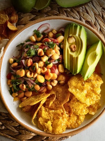 Overhead view of ceviche de chochos in a speckled bowl with lupini beans, red onion, cilantro, and corn nuts, alongside fanned avocado slices and crispy plantain chips.