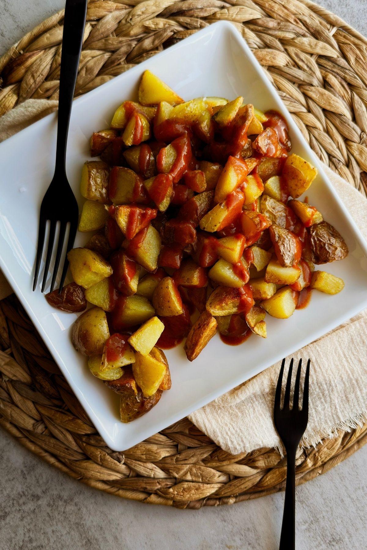 Patatas bravas on a square white plate, crispy potato chunks drizzled with smoky red bravas sauce, two black forks alongside.