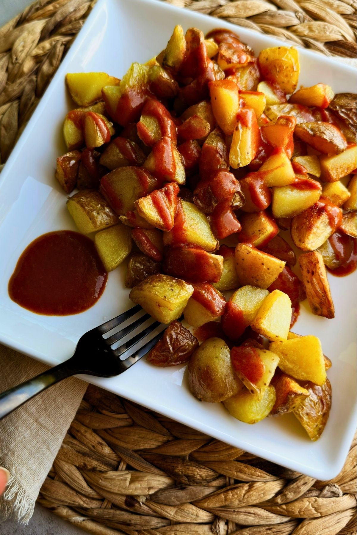 Close-up of patatas bravas on a white square plate, crispy potatoes drizzled with smoky red sauce, extra sauce pooled beside a fork.