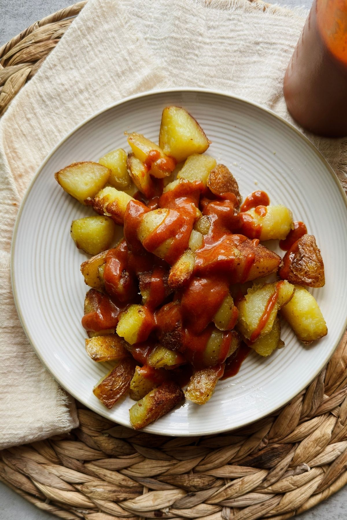 patatas bravas covered in bravas sauce on a round plate with a squeeze bottle of the sauce to the side.