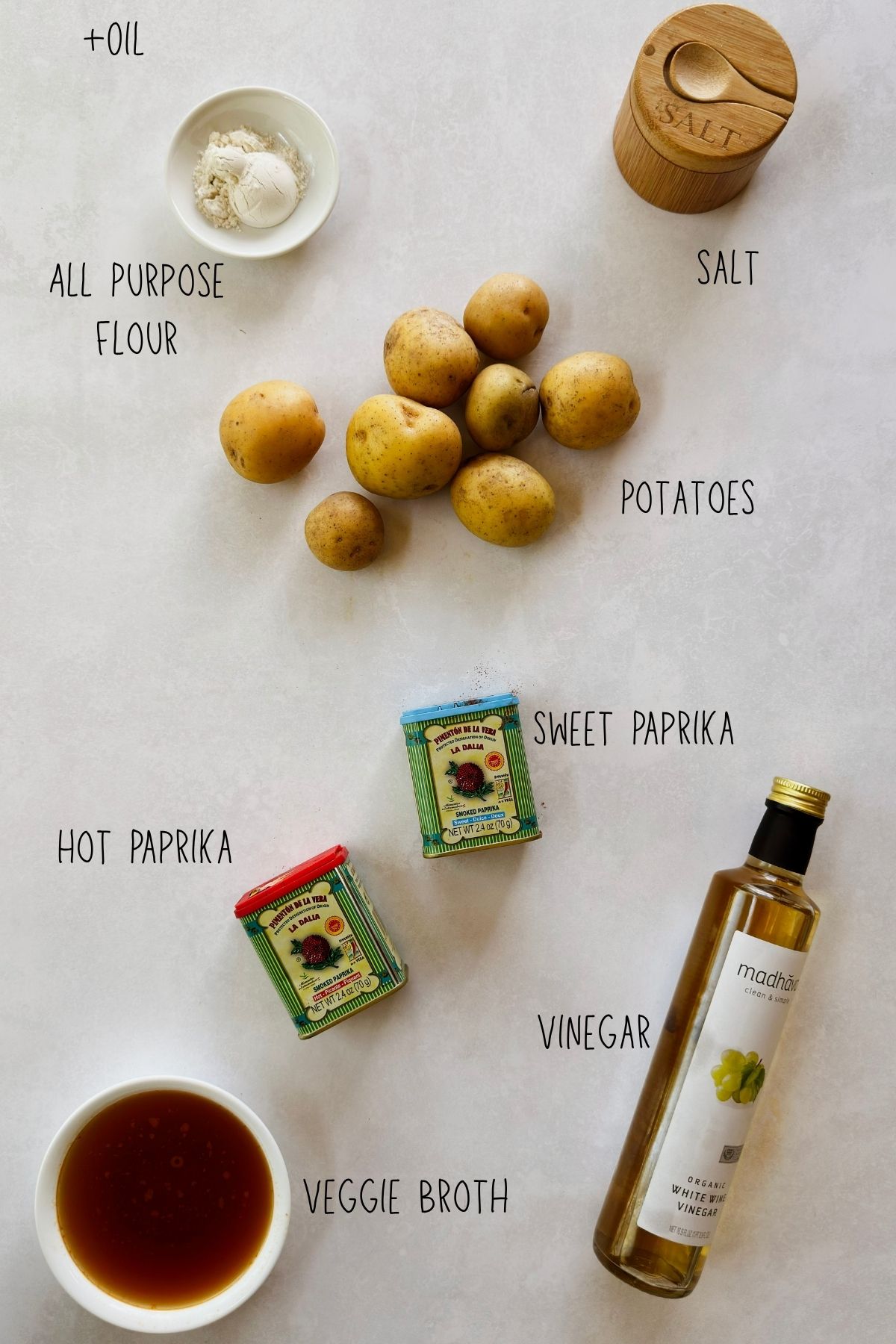 Labeled ingredients for patatas bravas on a white background: yellow potatoes, all-purpose flour, salt, sweet and hot smoked Spanish paprika tins, vegetable broth, and white wine vinegar.