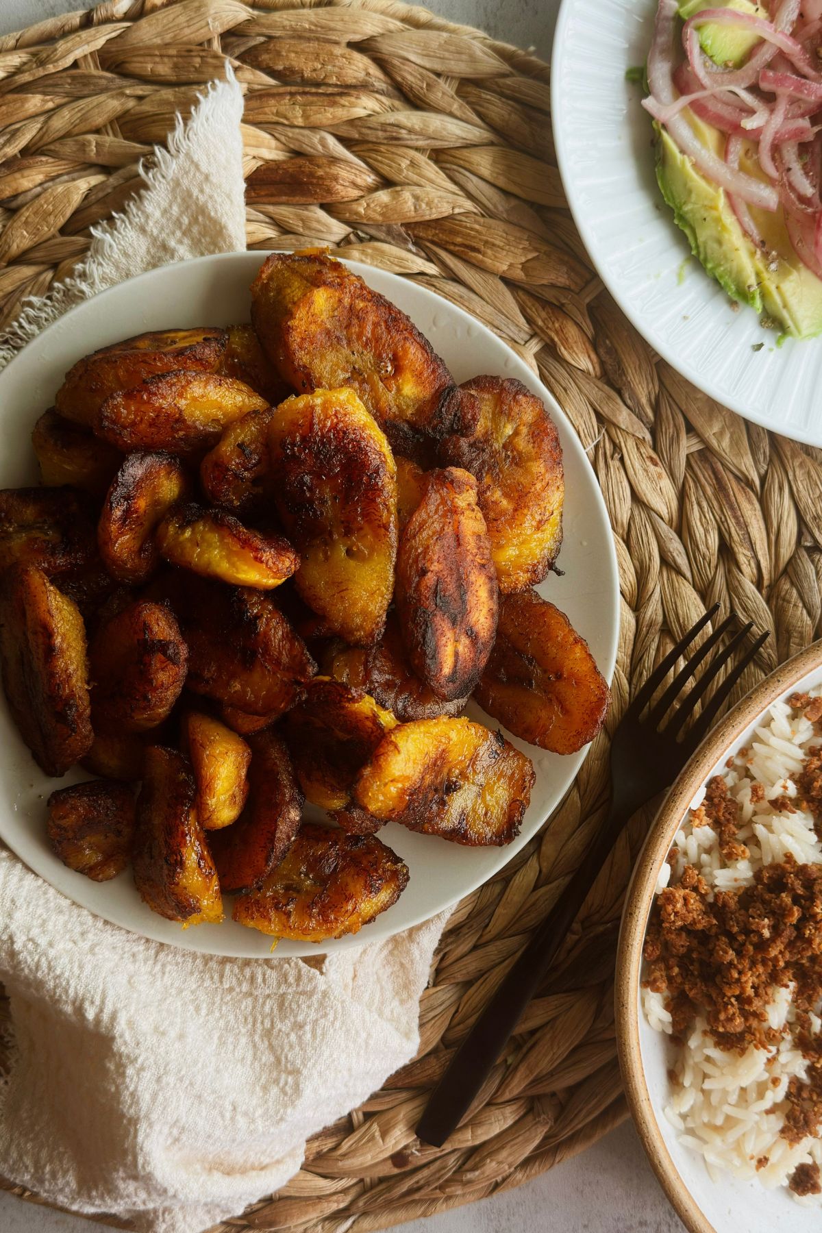 Plátanos maduros on a white plate with next to two plates. One with rice and vegan picadillo and the other with avocado, and pickled red onions