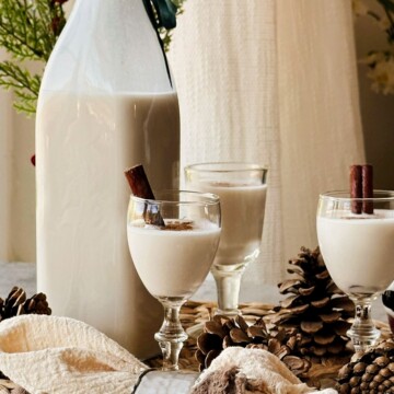 Tall swing-top bottle of creamy coquito beside three cinnamon-dusted cordial glasses, pinecones, and holiday greenery.