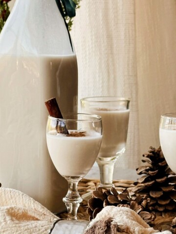 Tall swing-top bottle of creamy coquito beside three cinnamon-dusted cordial glasses, pinecones, and holiday greenery.