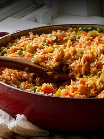 Red Dutch oven filled with vegan arroz con pollo, with wooden serving spoons resting in the golden rice.