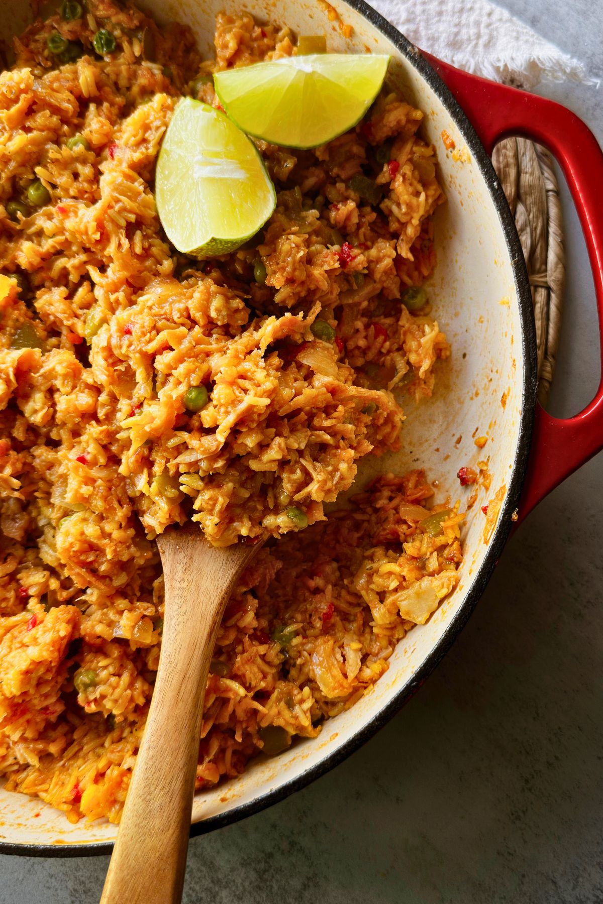 Close-up of saucy plant-based arroz con pollo in a red enameled pot, with lime wedges on top and a wooden spoon lifting a scoop of yellow rice.