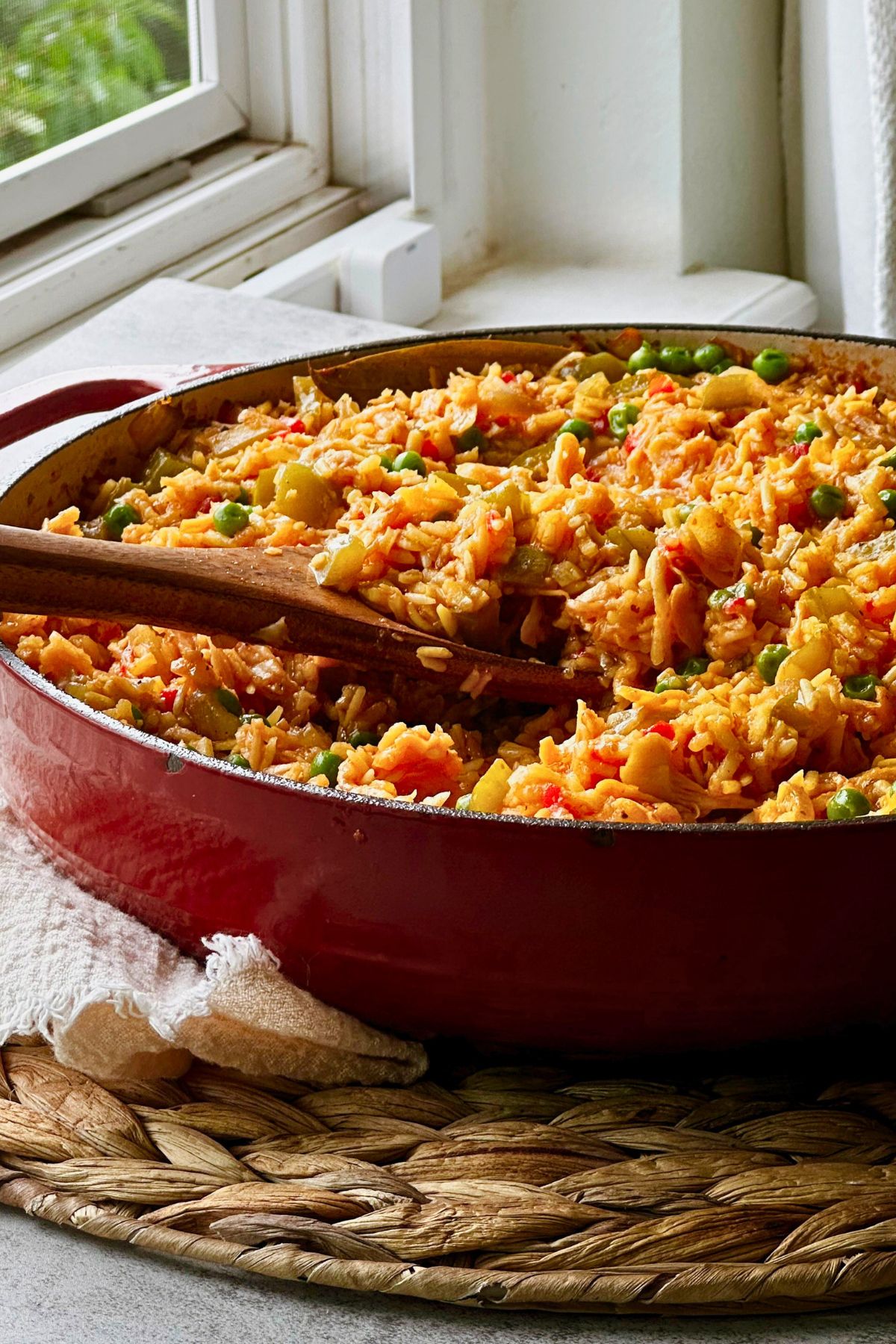 Red Dutch oven of vegan arroz con pollo by a sunny kitchen window, golden rice studded with green peppers, peas, and shredded vegan chicken, with a wooden spoon resting inside.