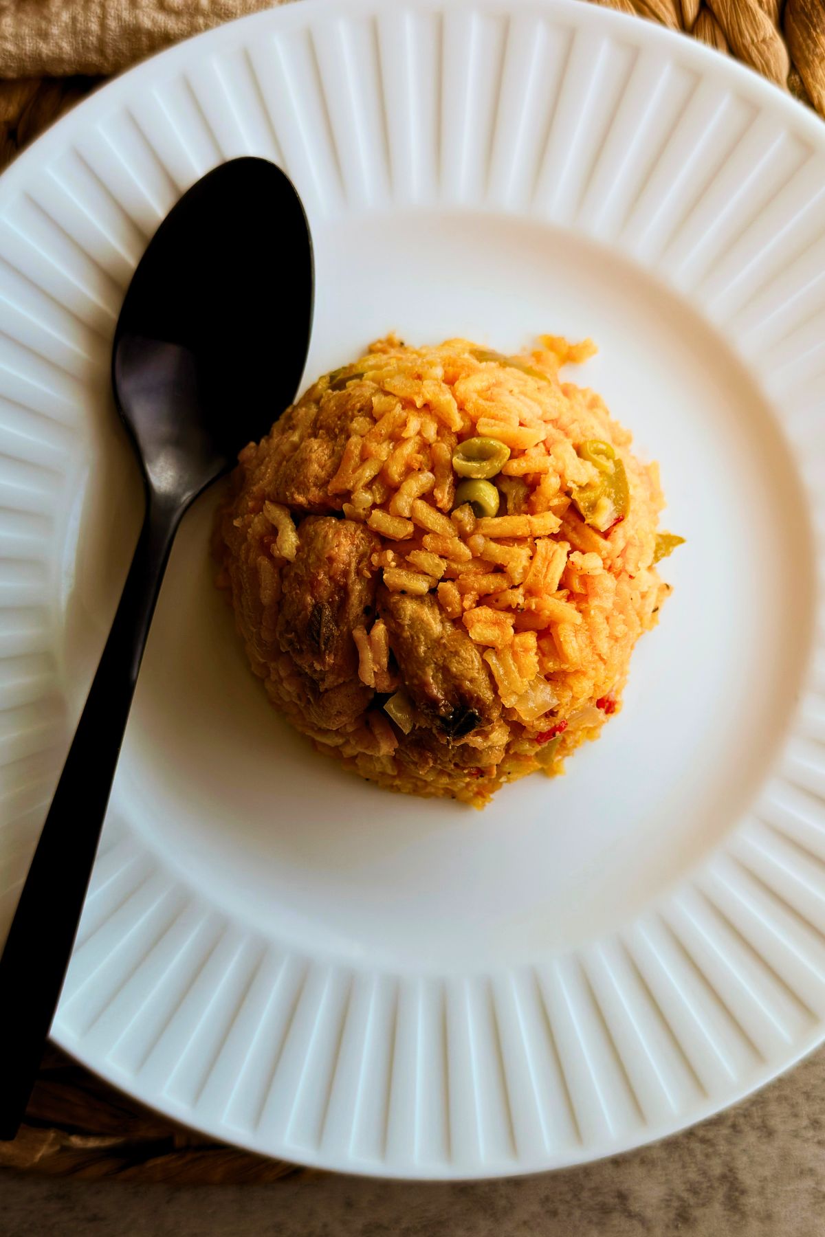 Rounded scoop of vegan chicken and yellow rice on a white plate with sliced green olives and a black spoon.