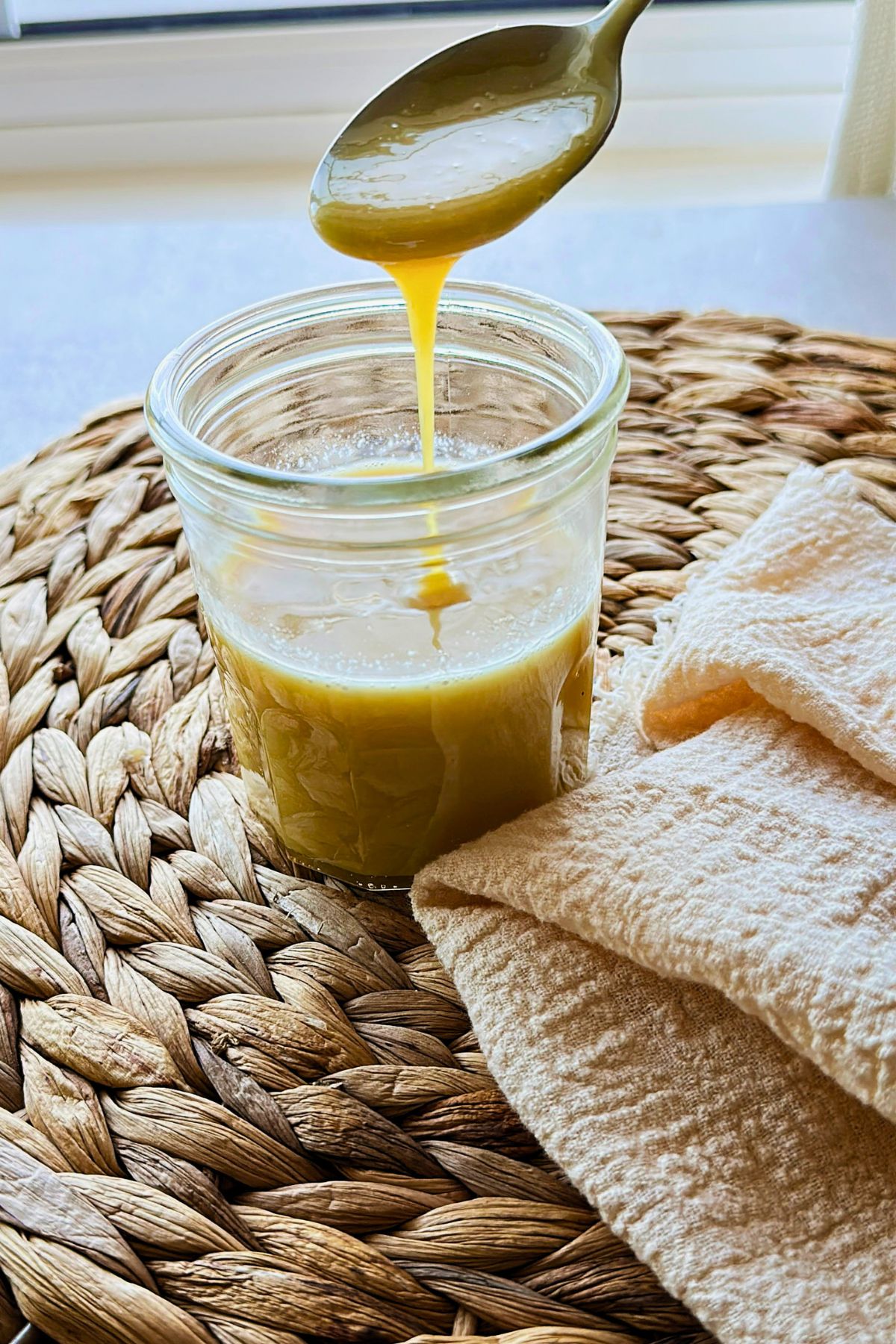 Thick golden vegan sweetened condensed milk drips from a spoon into a glass jar on a woven placemat beside a cream-colored towel.