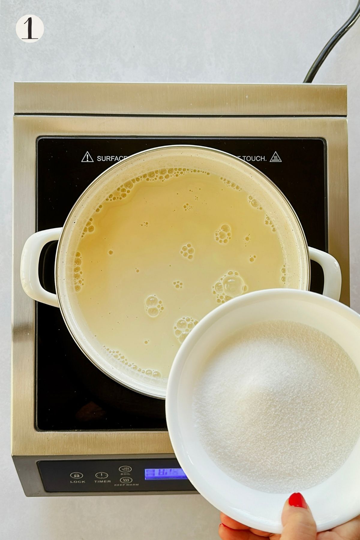 Step 1: A bowl of sugar is held over pale soy milk in a white saucepan on an induction burner.