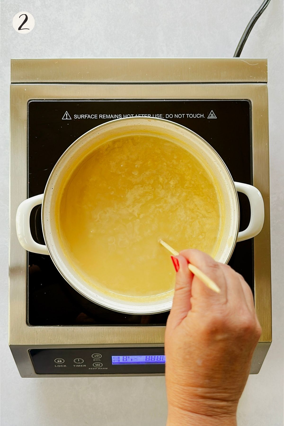 Step 2: Golden condensed soy milk simmers in a white saucepan while a hand stirs it with a wooden skewer.