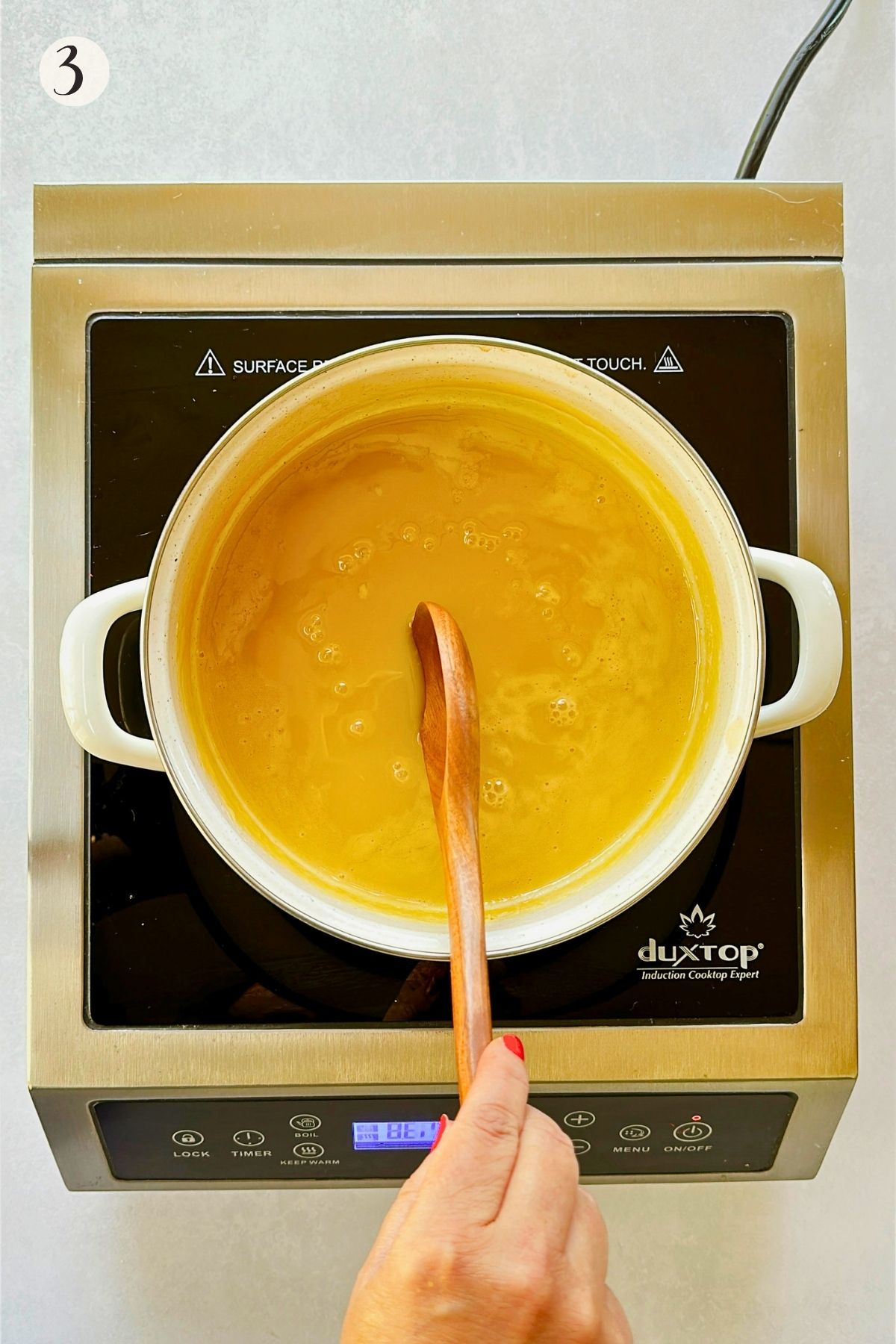 Step 3: Thickened golden condensed milk bubbles gently in a white saucepan while being stirred with a wooden spoon.