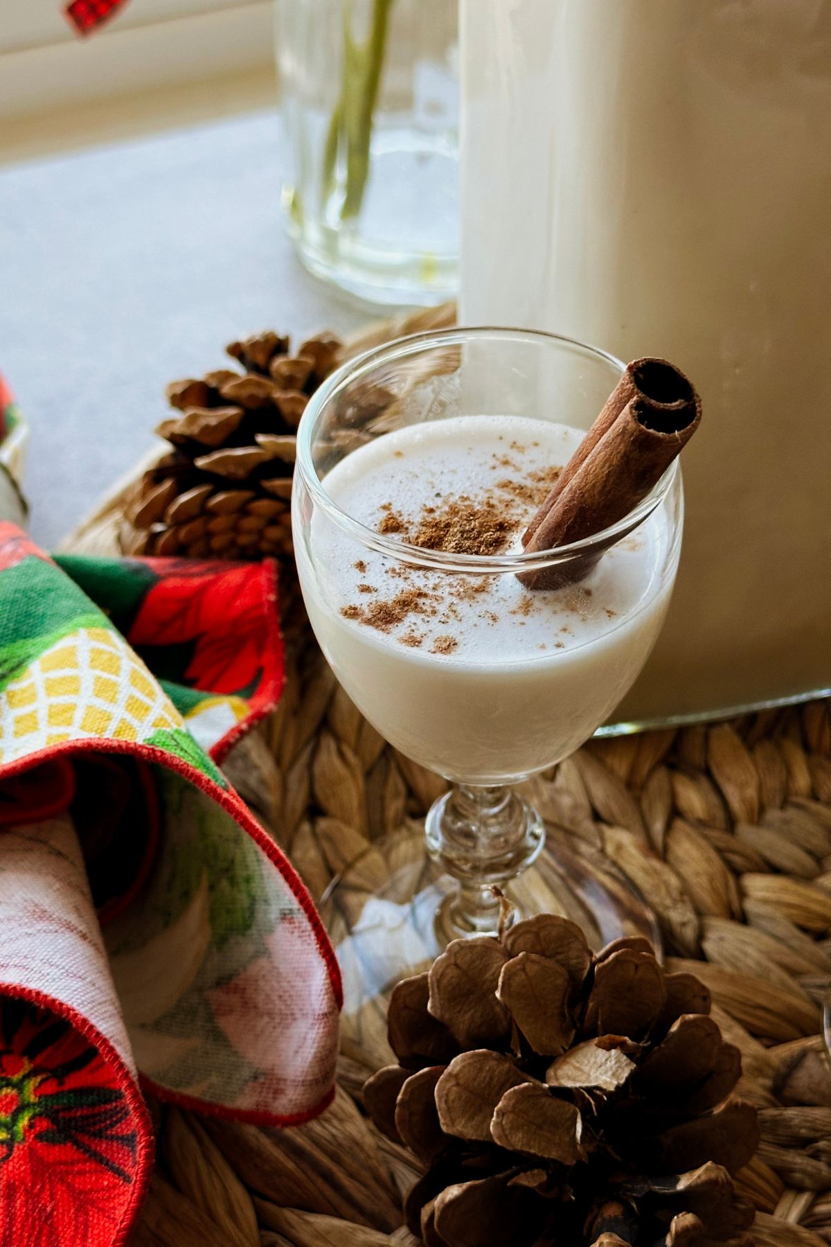Cinnamon-dusted coquito in a small stemmed glass with a cinnamon stick, beside a filled bottle, pinecones, and a red-and-green cloth.