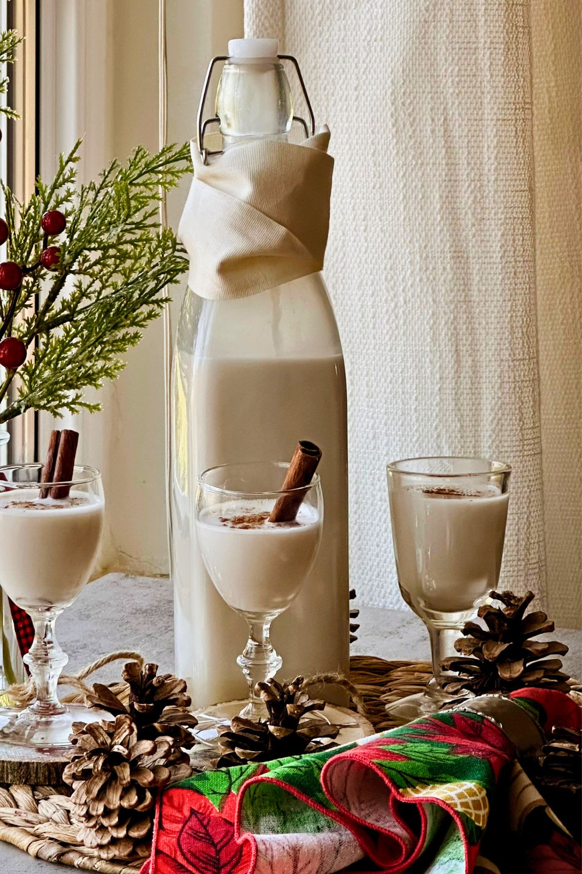 Bottle of creamy pale coquito with three cinnamon-dusted cordial glasses, cinnamon sticks, pinecones, and holiday greenery.